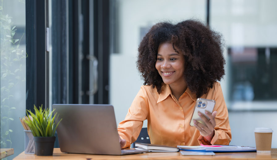 Woman with a phone looking at a laptop. 