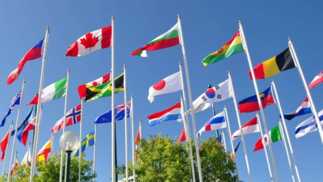 A vibrant display of international flags waves against a clear blue sky. The flags, representing various countries, including Canada, Japan, and South Korea, symbolize global unity and diversity. Green trees beneath add a natural touch to the colorful scene.